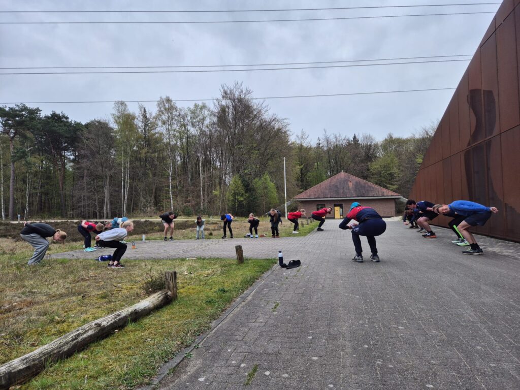 Droogtraining: leden doen schaatshoudingsoefeningen op een plein bij het bos